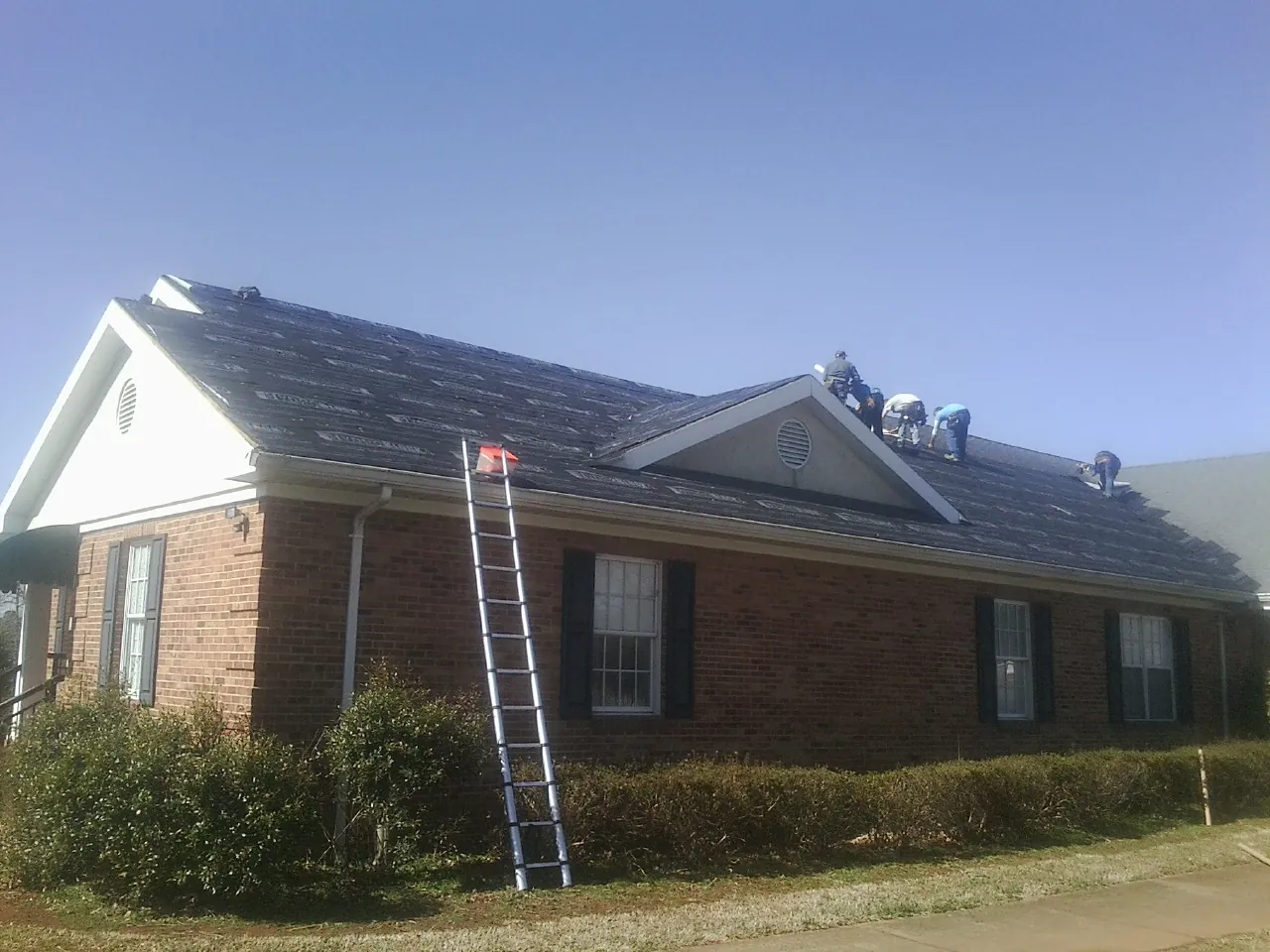 Crew tearing shingles off a brick ranch roof with a ladder set — Metal Roof Repair work by our crew in Tatums